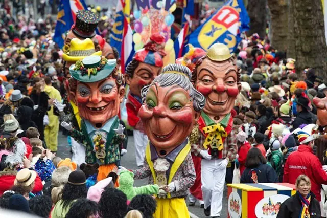 A carnival parade of comic figures with huge heads crosses a street overflowed with a cheerful crowd.