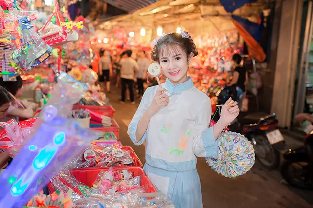 A girl standing in front of a bench of an open-air market and holding ornaments of the Mid-Autumn Festival.