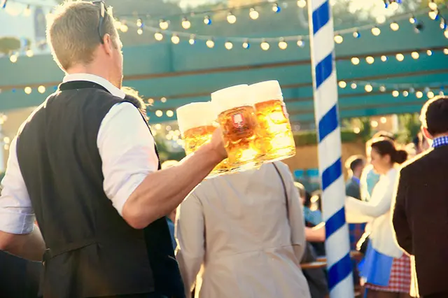A waiter is serving large glasses of beer in a crowded open-air pub during Octoberfest.