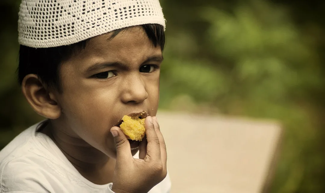 A little kid wearing a kufi prayer cup (taqiyah) is eating with his hand a piece of bread.